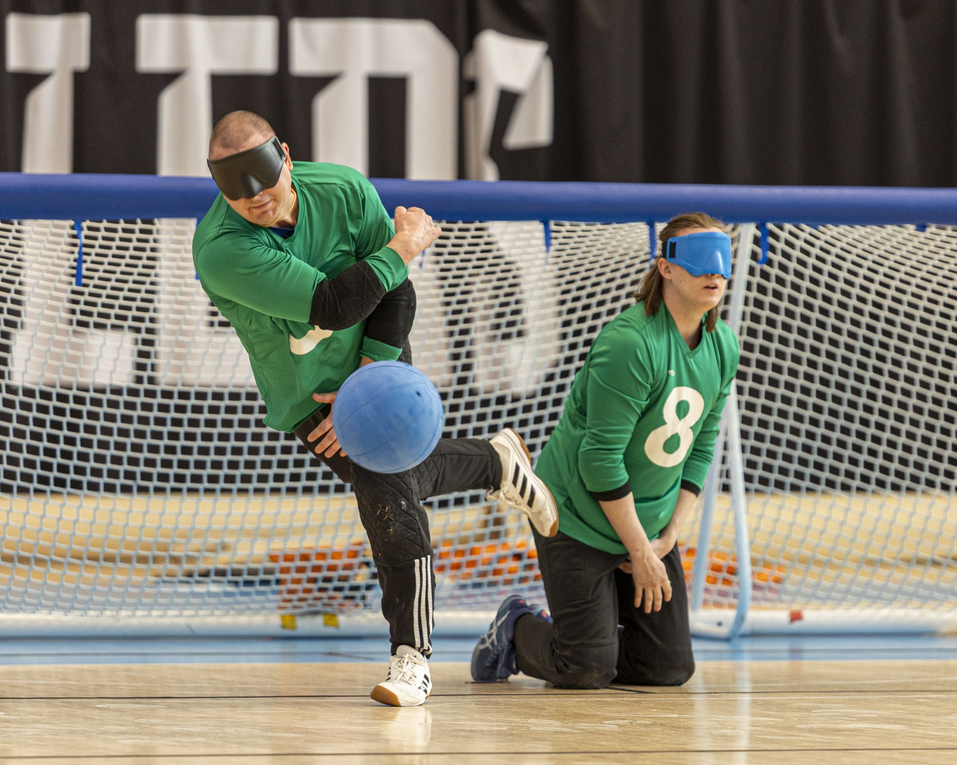 Posio heittää maalin edessä pystyssä palloa, joka lentää kohti kameraa. Linnas hänen takanaan maassa puolustusvalmiudessa.