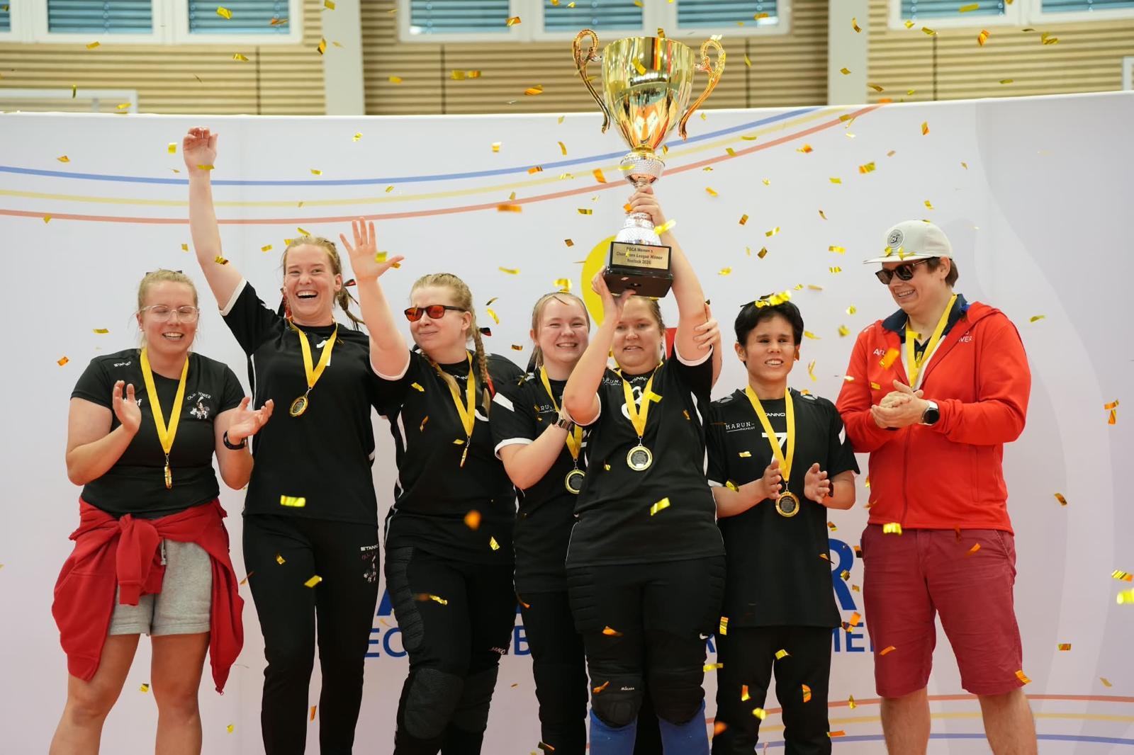 Naisten joukkue tuulettamassa pokaalin ja mitaleiden kanssa konfettisateessa.