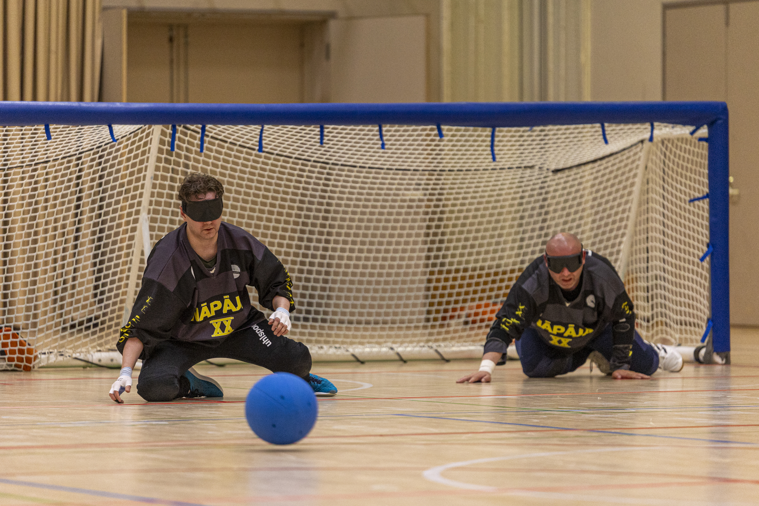 Maalipallo pelikuva. Kaksi Näpäjä XX: pelaajaa mustissa pelipaidoissa torjumassa kohti tulevaa palloa.