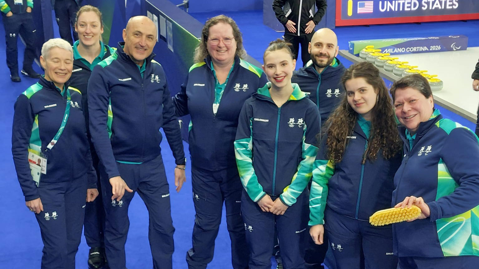 Kaisla Heikkinen yhteiskuvassa muiden curlingin jäätiimin toimitsijoiden kanssa. Päällään heillä kisavarustus. Kaikilla hymy kasvoilla. Taustalla curling-kenttä.