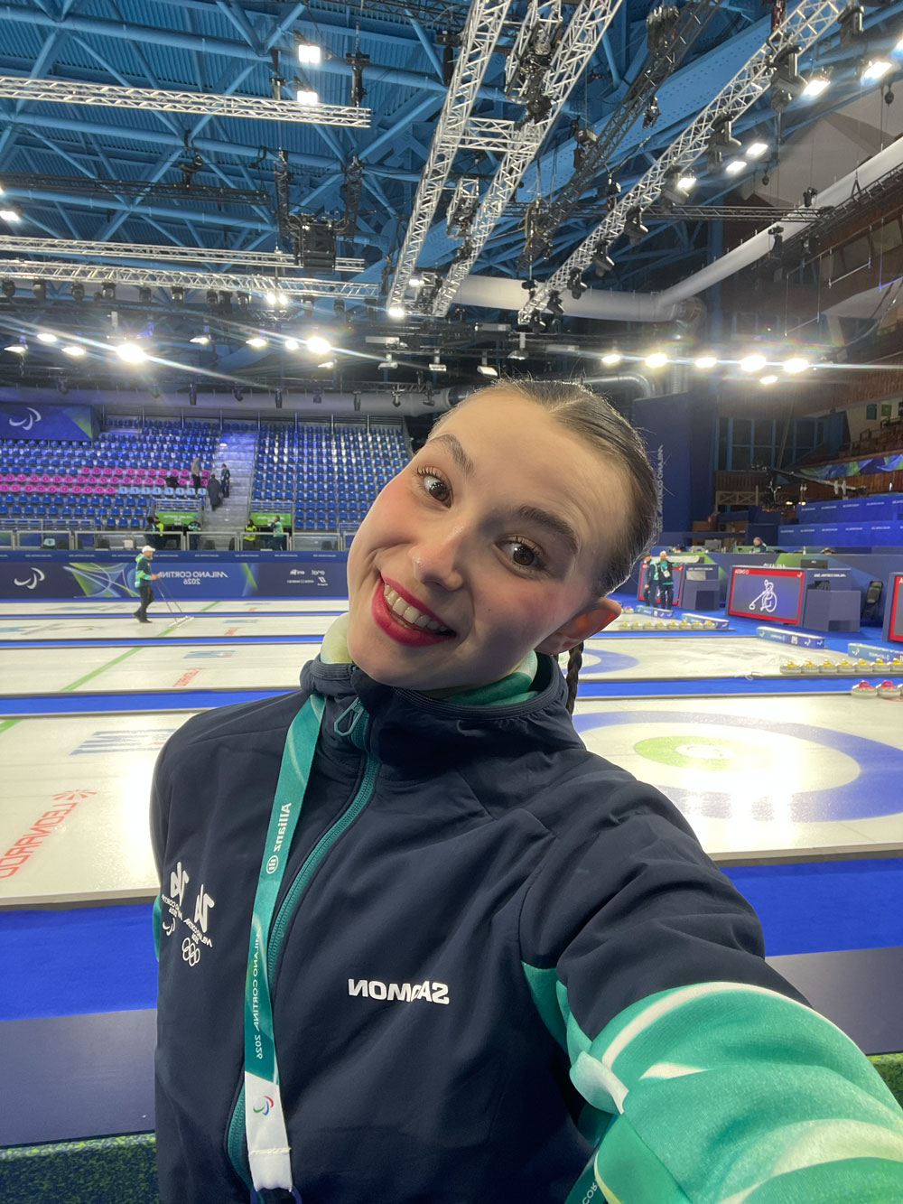 Kaisla Heikkinen selfie-kuvassa curling-kenttää vasten. Päällään kisavarustus, ja kasvoillaan hammashymy.