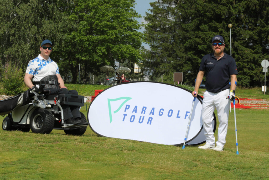 Kaksi golfaria poseeraamassa kameralle paragolf tour kyltin vieressä. Toinen käyttää paragolfer laitetta ja toinen kyynärsauvoja.