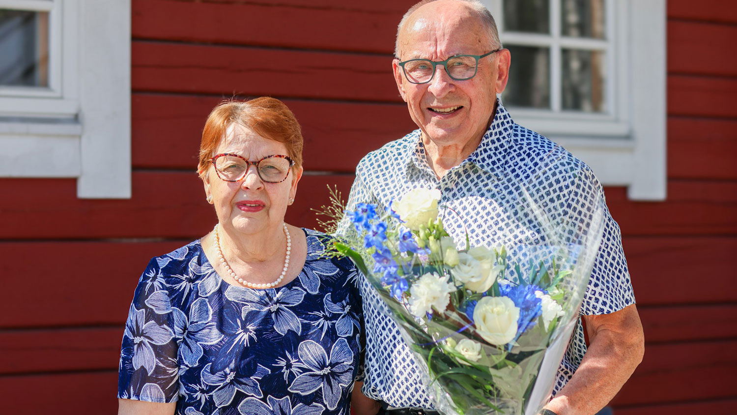 Pertti Sankilampi kukkakimppu kädessä hymyilemässä yhdessä vaimonsa Sirkan kanssa punaisen puurakennuksen edessä aurinkoisessa säässä.