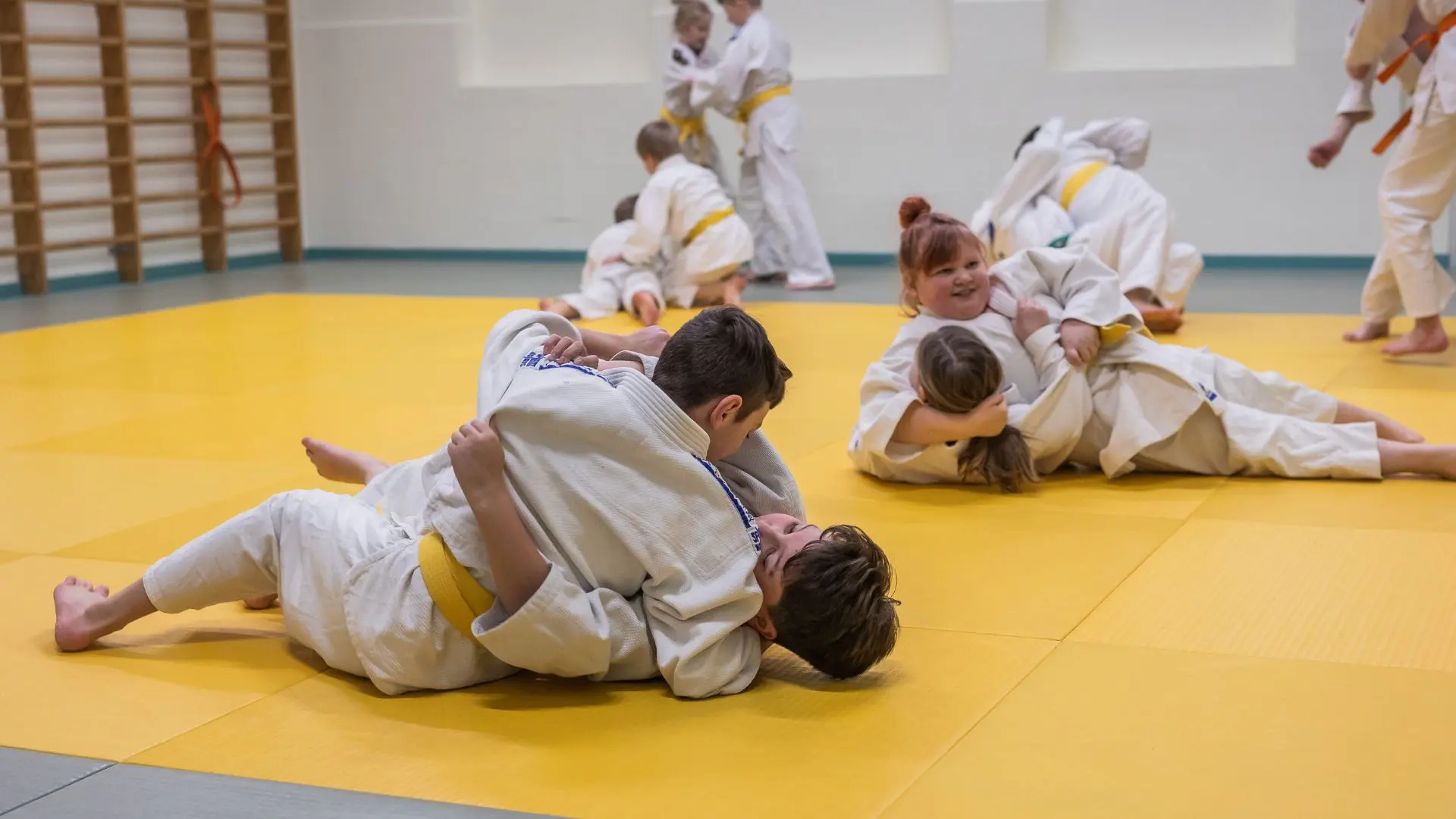 Henkilöitä judo-treeneissä keltaisella tatamilla.