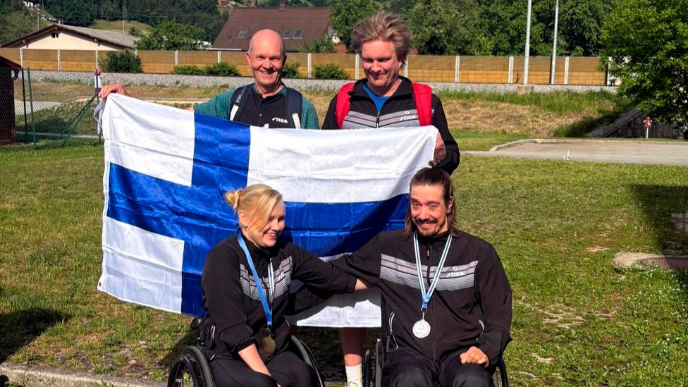 Aino Tapola ja Timo Natunen valmentajineen poseeraamassa kameralle mitalit kaulassaan, Suomen lippu taustallaan.
