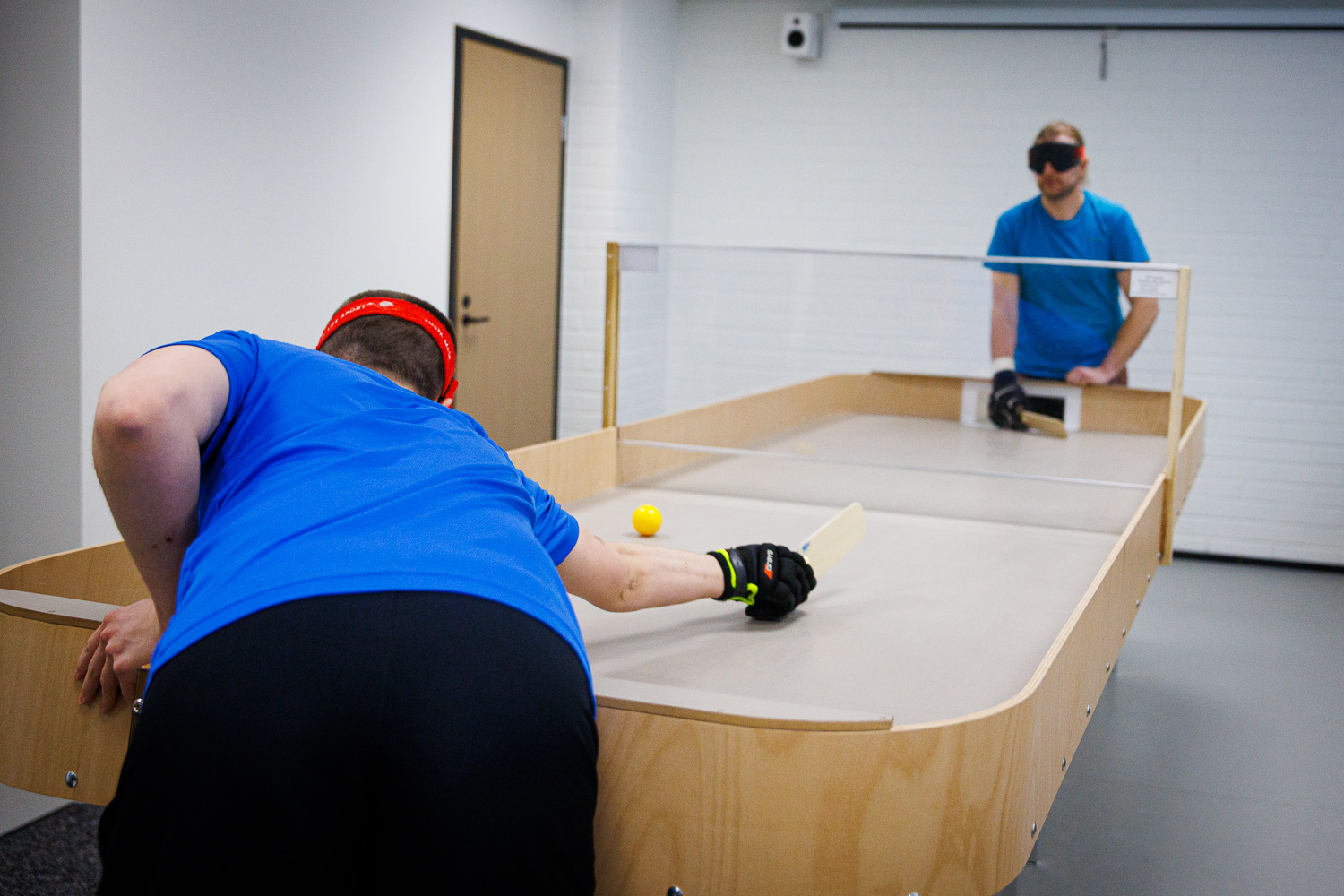 Etualalla Jari Linnavuori selkä kameraan tähtäämässä sokkopingispalloon. Blurrattuna taustalla, kasvot kameraan Väinö Rihti, myöskin peliareenalla.