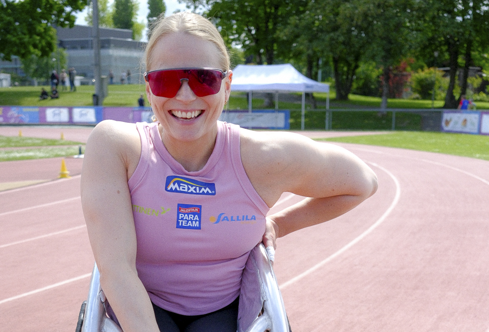 Amanda Kotaja hymyilemässä kameralle aurinkolasit kasvoillaan, pinkki toppi päällään, jossa sponsorien logoja. Lokaationa yleisurheilukenttä ulkona.