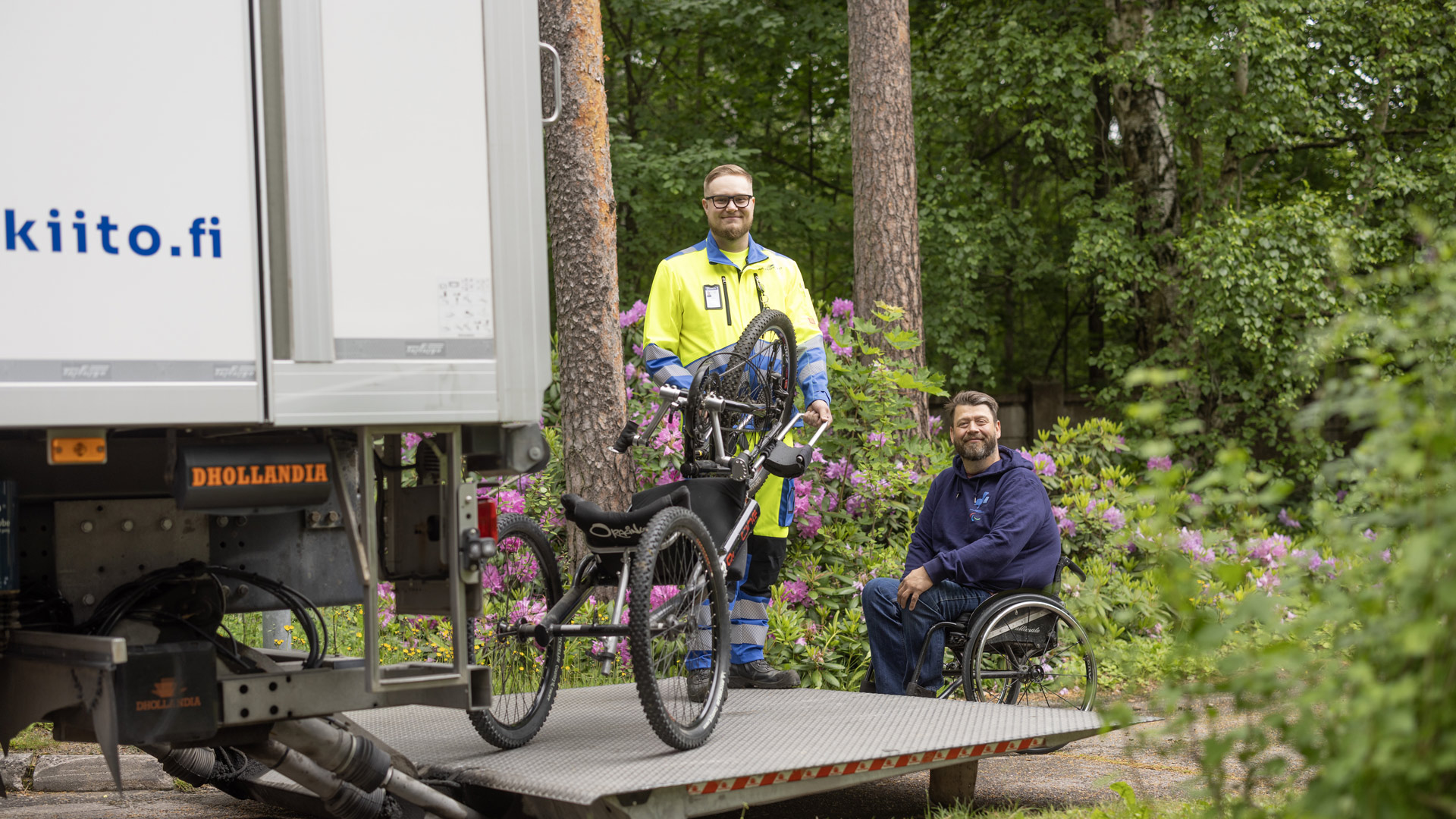 Kaukokiidon työntekijä ja välineen vuokrannut pyörätuolia käyttävä henkilö vierekkäin rekan vieressä, josta otetaan ulos apuvälinettä.