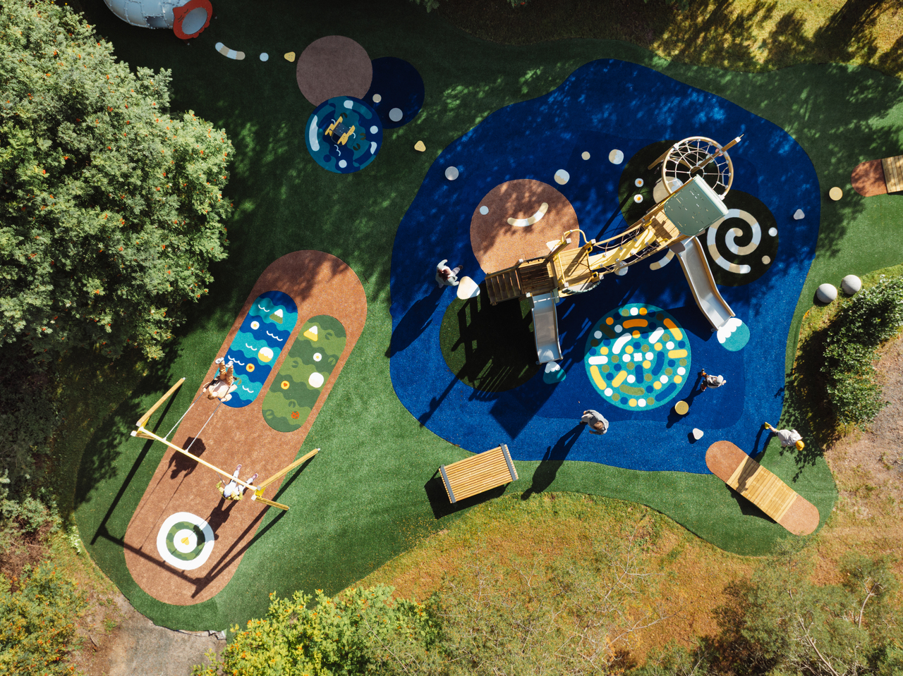 Lappsetin Prime-malliston kesäinen havainnekuva: ylhäältäpäin kuvattu värikäs leikkipuisto, jossa tilavia keinuja, moniosainen kiipeilyteline liukumäkineen ja maahan nurmikkoon maalattuja ratoja.