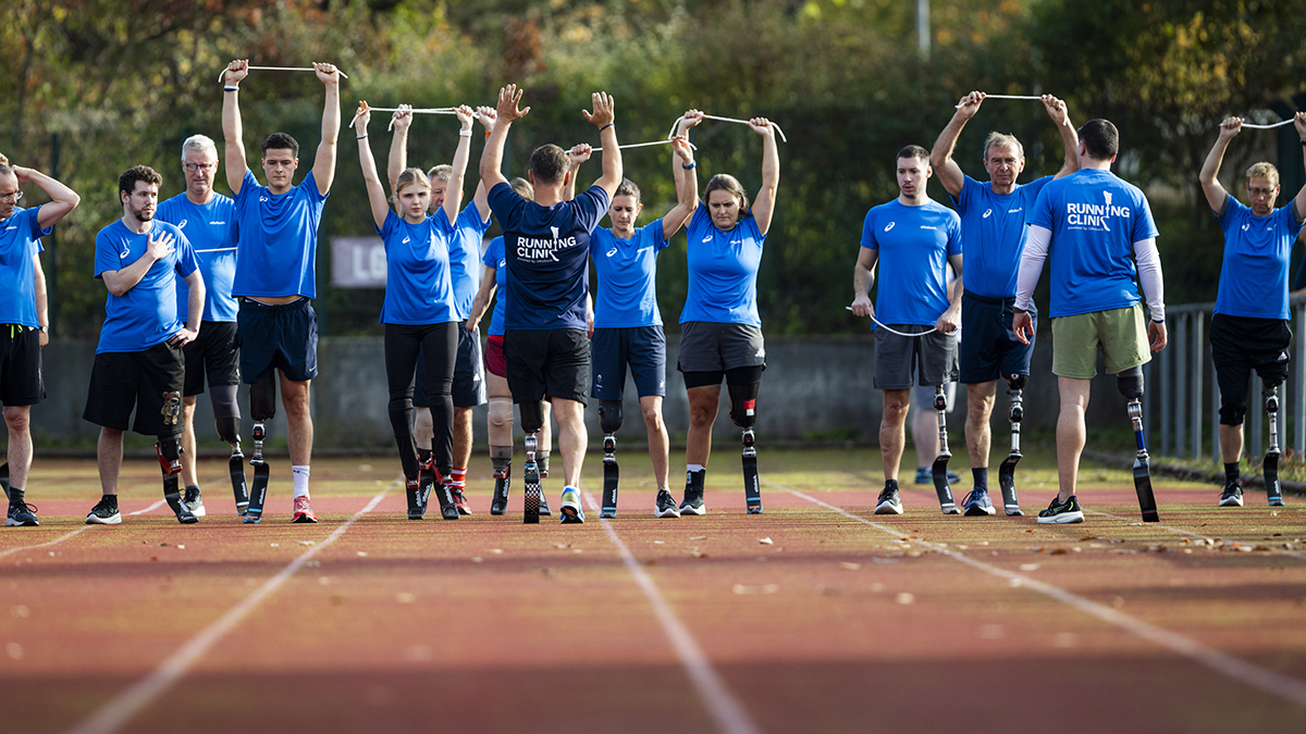 Eri ikäisiä ihmisiä venyttelemässä yleisurheilukentän juoksuradan lähtöviivalla, kaikilla toisessa jalassa juoksuproteesi. Selin kameraan ohjaajia, joiden paidan selässä näkyy teksti "Running Clinic".