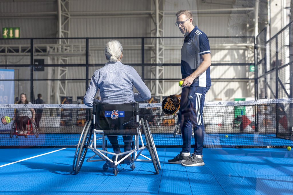 Pyörätuolia käyttävä henkilö pelaamassa tennistä, ohjaaja vierellä.