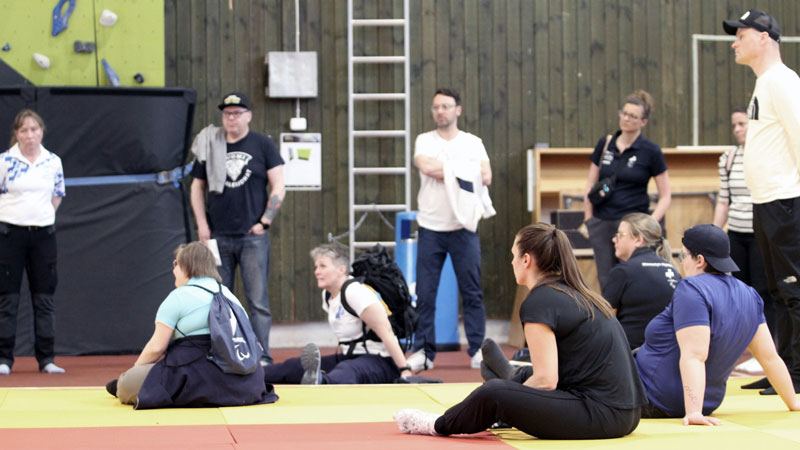 Henkilöitä istumassa ja seisomassa liikuntahallin lattialla, seuraamassa koulutusta.