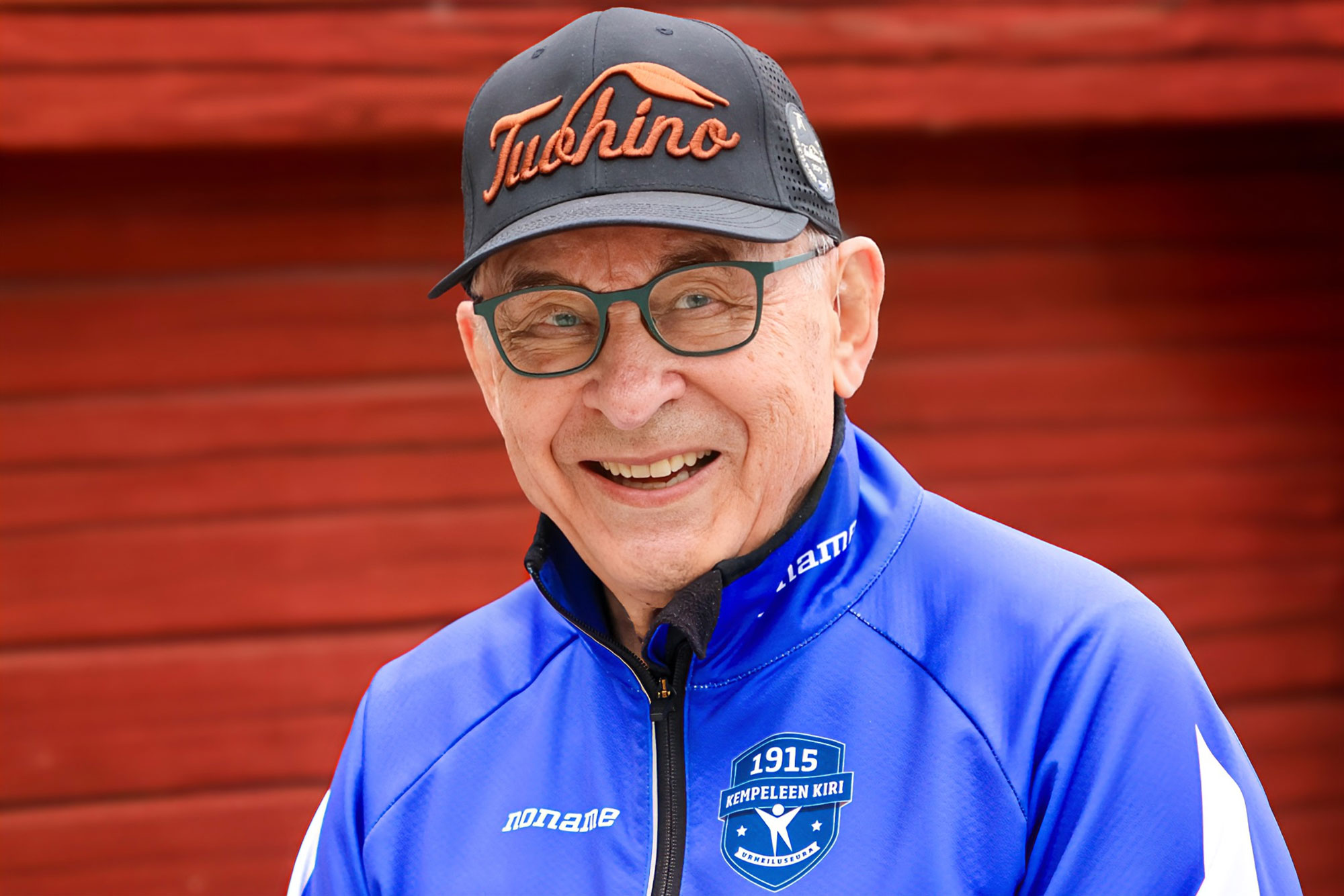 Pertti Sankilampi hymyilemässä kameralle punaisen puurakennuksen edessä.
