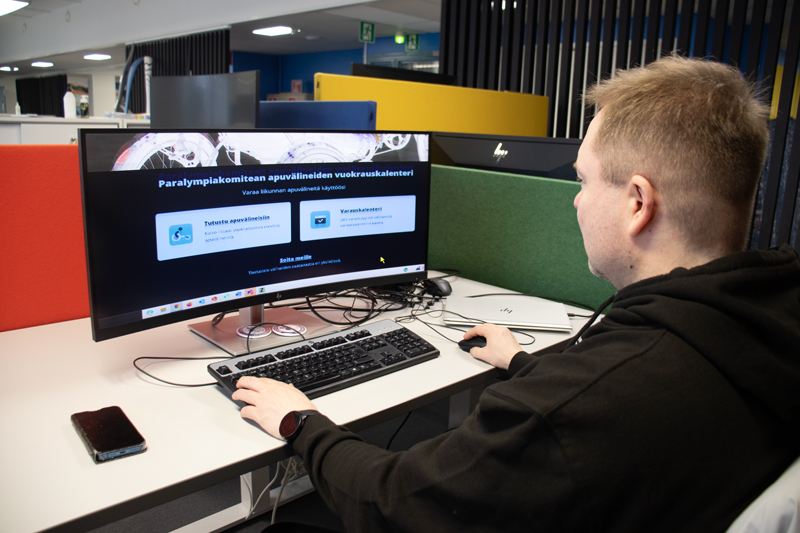 Näkövammainen henkilö käyttämässä Paralympiakomitean apuvälinetoiminnan varauskalenteria tietokoneella avokonttorissa.