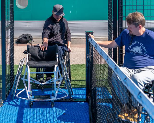 Pyörätuolia käyttävä henkilö tulossa sisään esteettömälle padelkentälle, erillistä pelituolia edessään työntäen. Toinen pyörätuolia käyttävä henkilö odottaa keskiverkon toisella puolella.