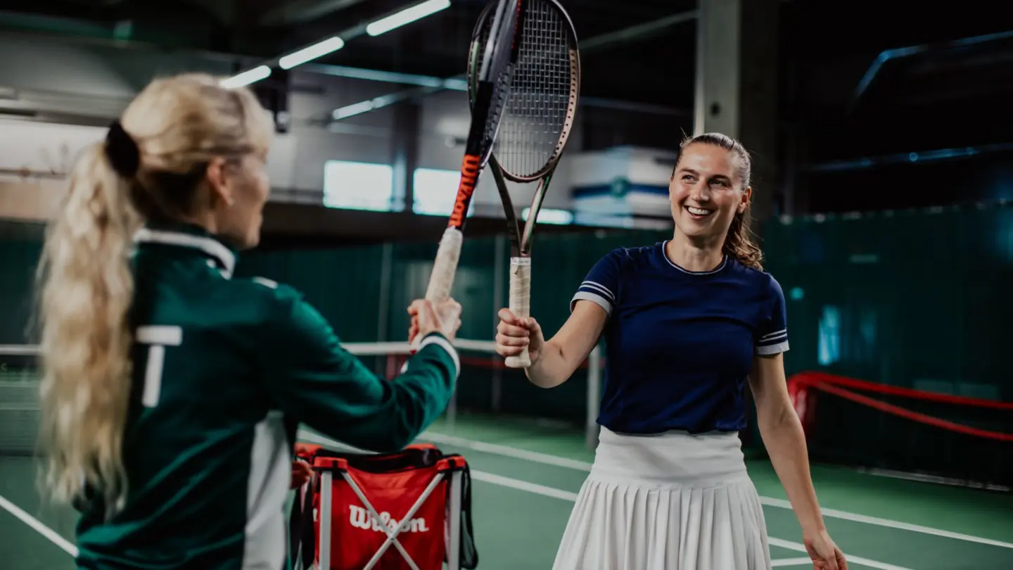 Kaksi iloista naistennispelaajaa kopauttamassa mailojaan yhteen.