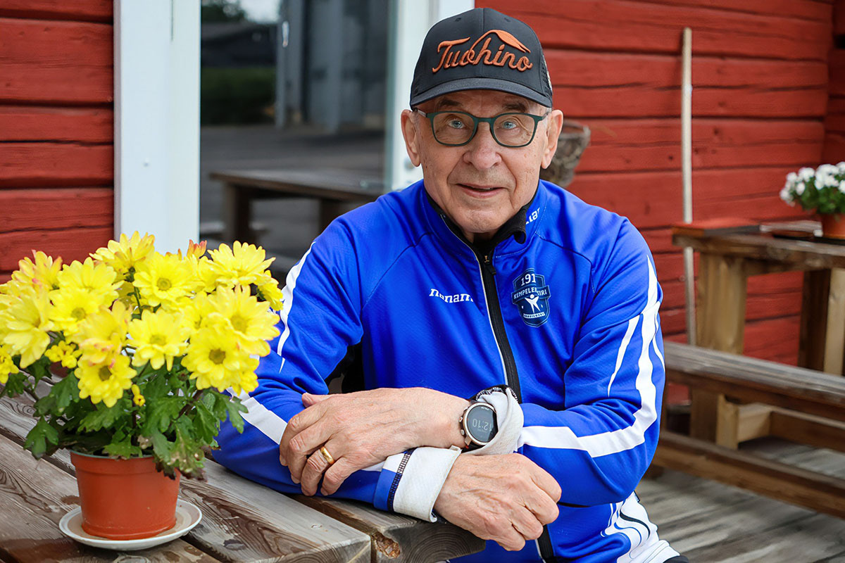 Pertti Sankilampi sinisessä verkkatakissa ja mustassa lippiksessä poseeraa kameralle punaseinäisen aittarakennuksen edessä ulkona. Pöydän ääressä hänen edessään keltaisia kukkia ruukussa.
