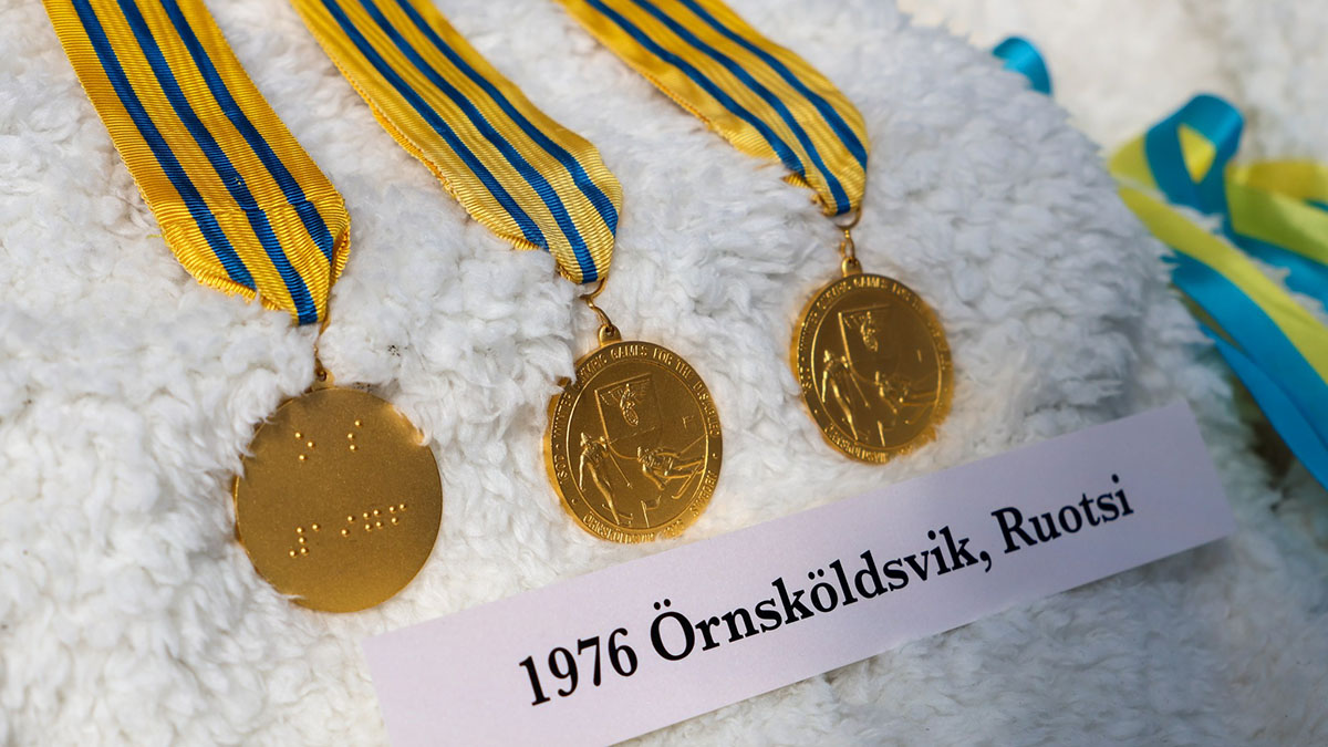 Kolme kultaista mitalia keltasinisissä kaulanauhoissa aseteltuna riviin pehmustetulla alustalle. Alla paperinen tekstikyltti: "1976 Örnsköldsvik, Ruotsi".