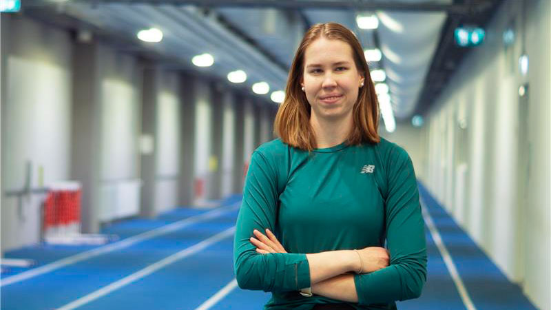 Iida Lounela hymyilemässä kameralle sisäjuoksuradalla. Päällään vihreä, pitkähihainen urheilupaita. Kädet ristissä.