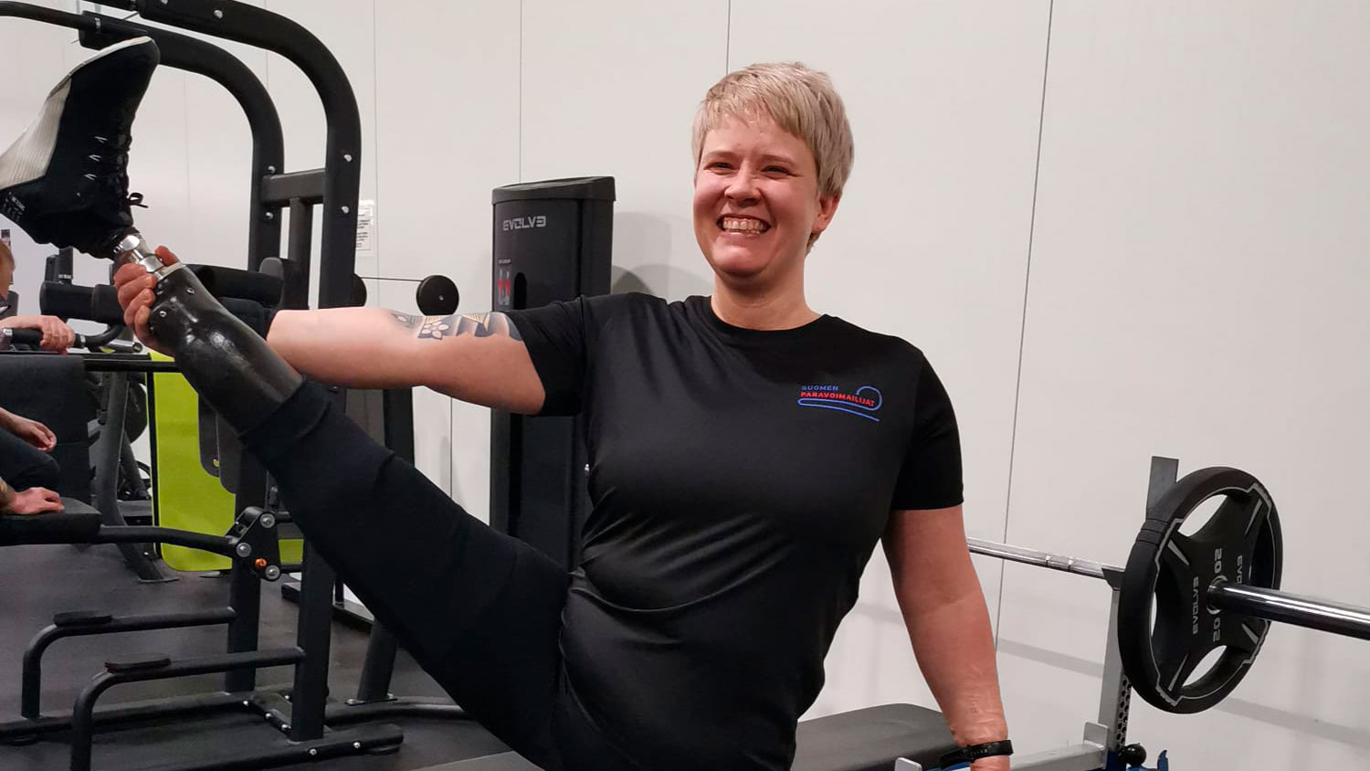 Nelli Kinnunen hymyilemässä iloisena kameralle nostaen proteesijalkaansa ylös sivulle kädellä avustaen.