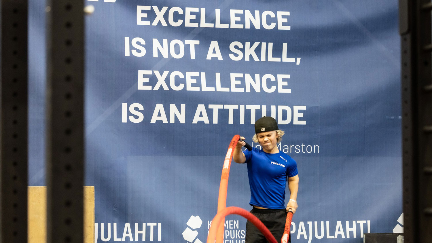 Aukusti Paakkala treenaamassa köysien kanssa. Taustalla sininen lakana, jossa valkoinen teksti capslockilla: Excellence is not a skill, excellence is an attitude.