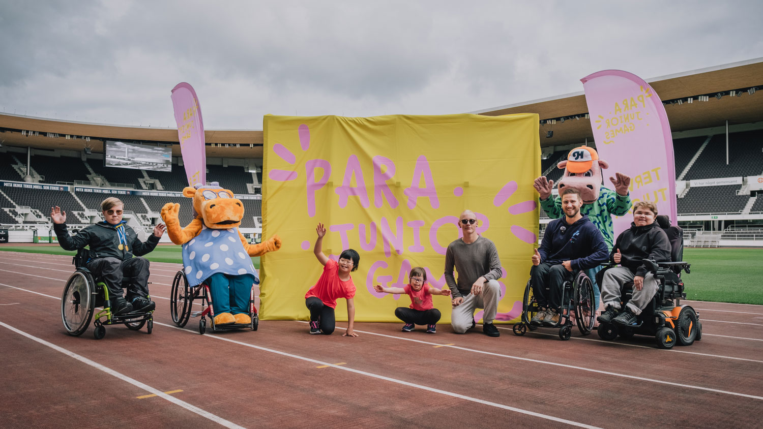 Iloisia lapsia ja nuoria yhdessä OP:n hippojen kanssa. Toinen hipoista käyttää pyörätuolia. Lokaationa Olympiastadion Helsingissä. Taustalla Para Junior Games -lakana ja beach flageja.
